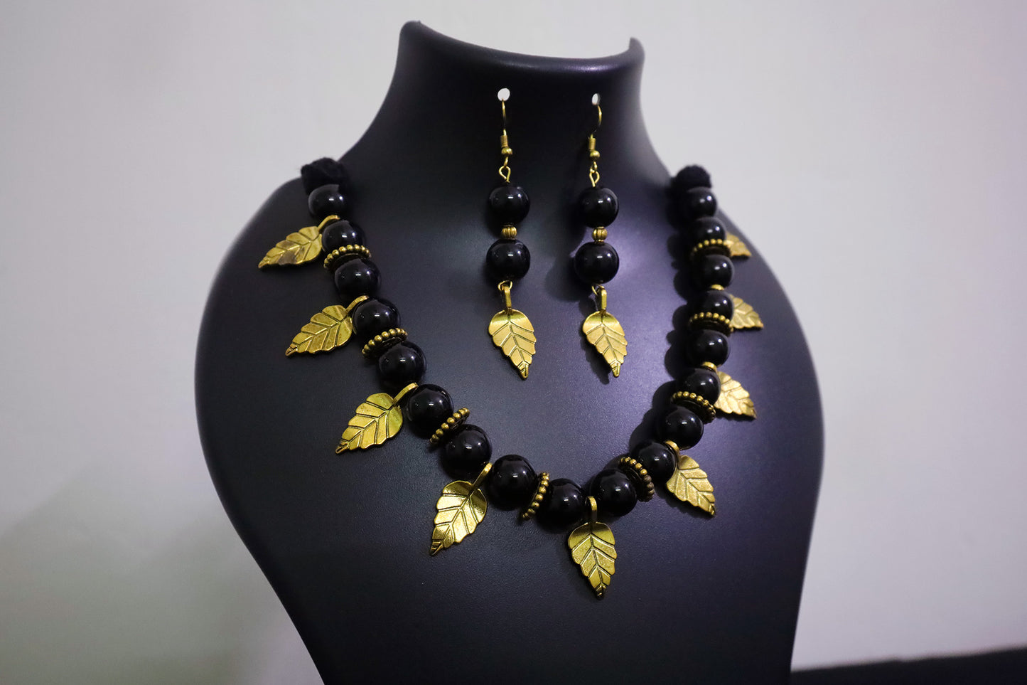 Closer view of handmade black beads and golden leaf charm necklace with earrings on a black mannequin.