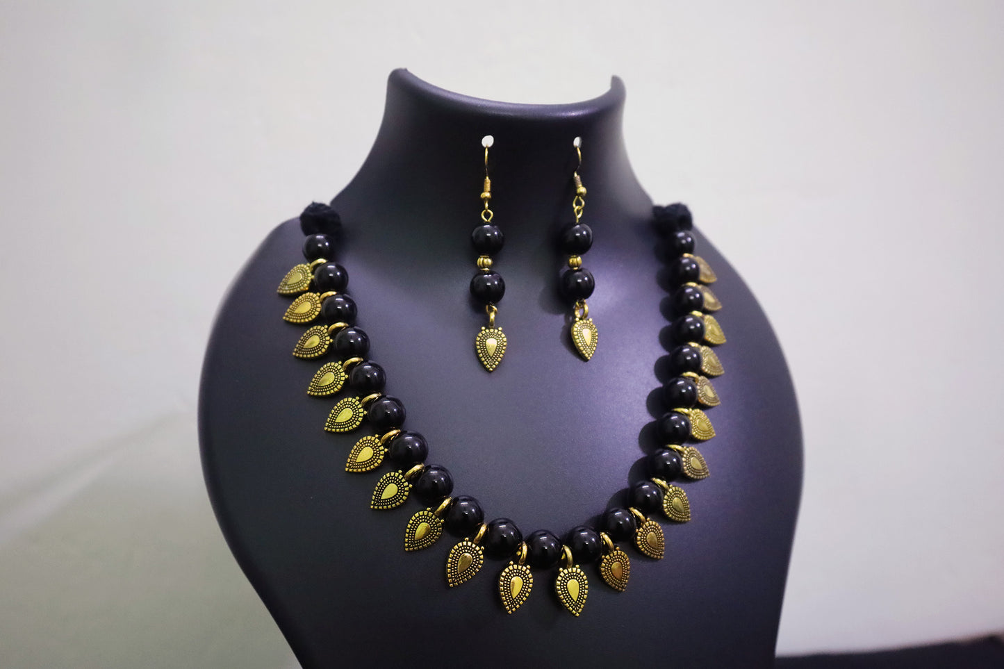 Black glass beaded with golden leaf charm necklace and earrings set on a black mannequin bust against a white background