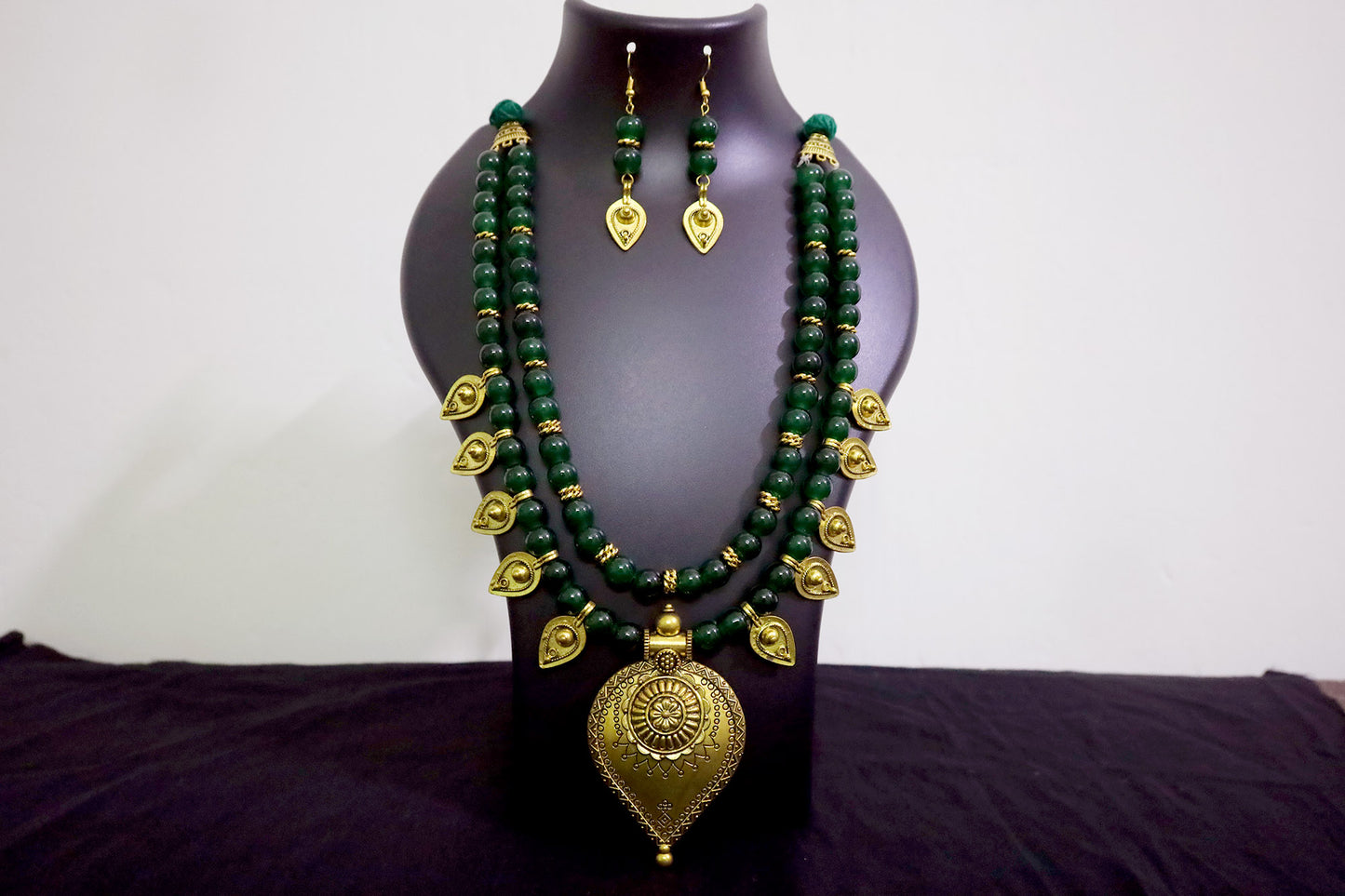 Closer look of dark green glass beads and golden drop pendant necklace with earrings on black mannequin.