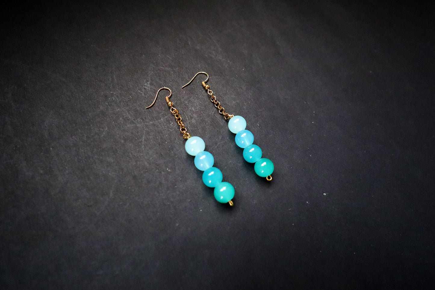 Angled view of Pair of Turquoise handcrafted Gradient Glass Bead & Golden Chain Dangle Earrings on black fabric background