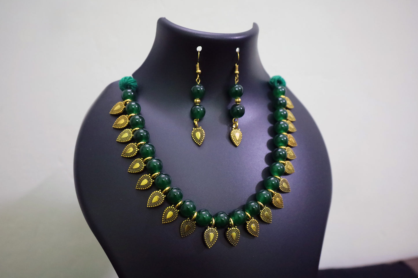 Green glass beaded with golden leaf charm necklace and earrings set on a black mannequin bust against a white background
