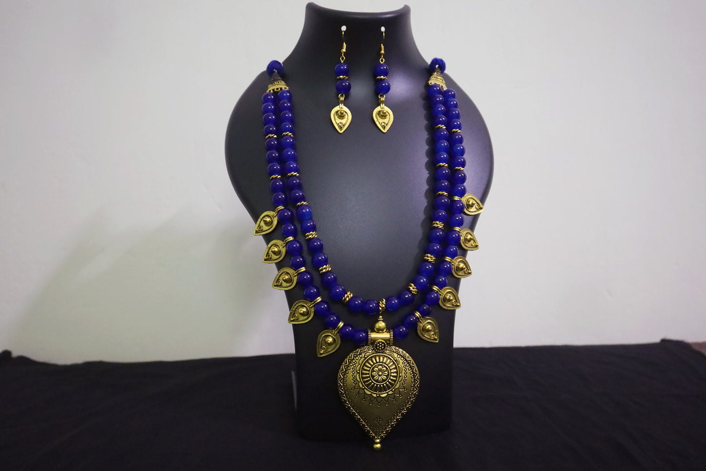 Closer look of blue glass beads and golden drop pendant necklace with earrings on black mannequin.