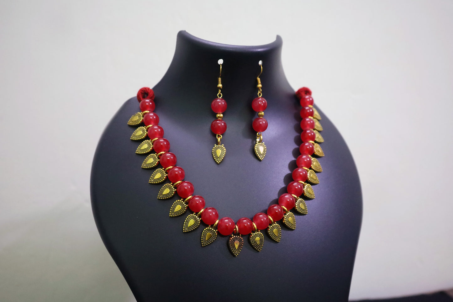 Red glass beaded with golden leaf charm necklace and earrings set on a black mannequin bust against a white background