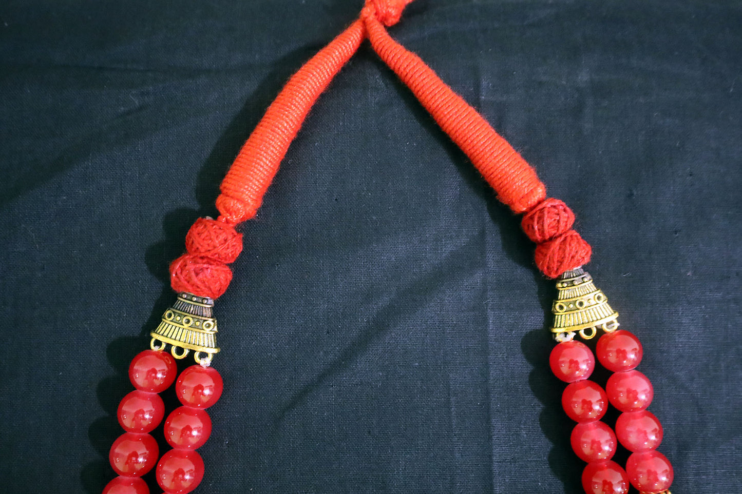Closer top section of red necklace with glass beads and golden drop pendant on black fabric.