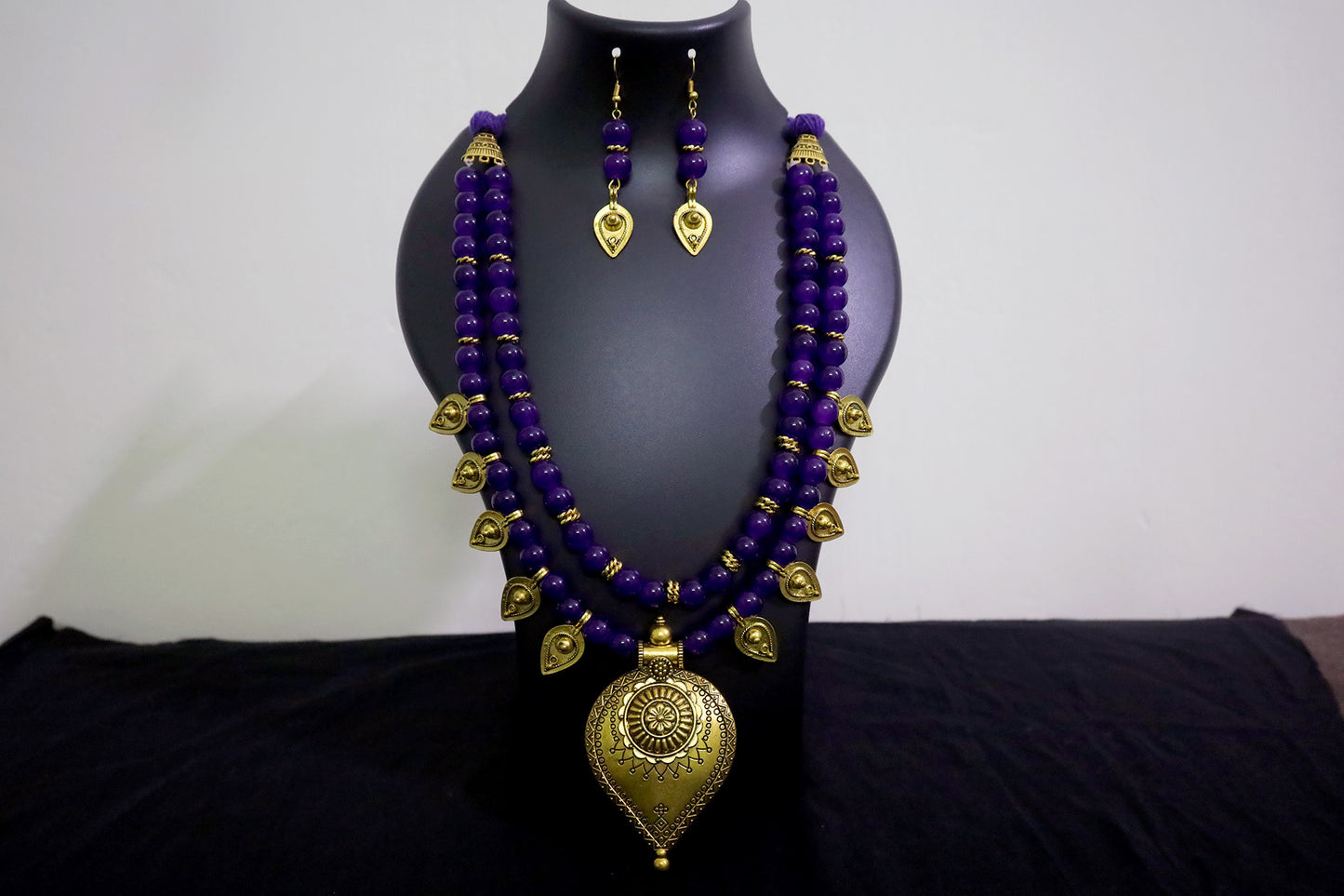 Closer look of violet glass beads and golden drop pendant necklace with earrings on black mannequin.