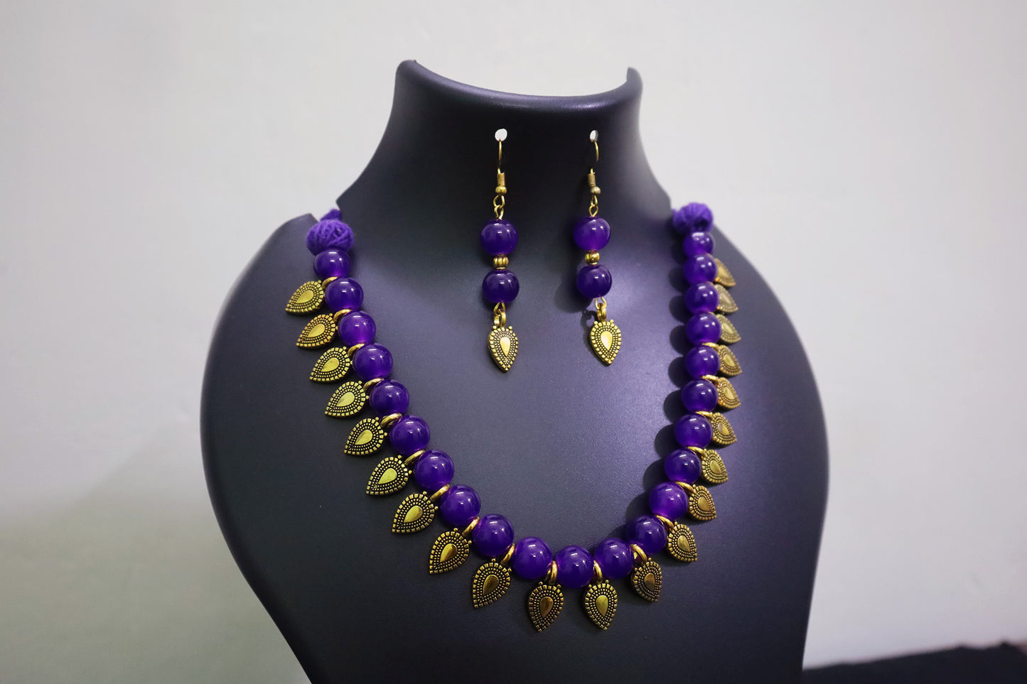 Violet glass beaded with golden leaf charm necklace and earrings set on a black mannequin bust against a white background