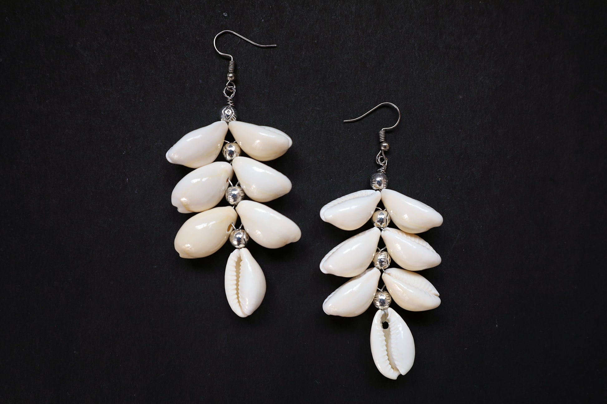 Top view of pair of Cowrie Seashell & Metal Ball Dangle Earrings on black fabric background