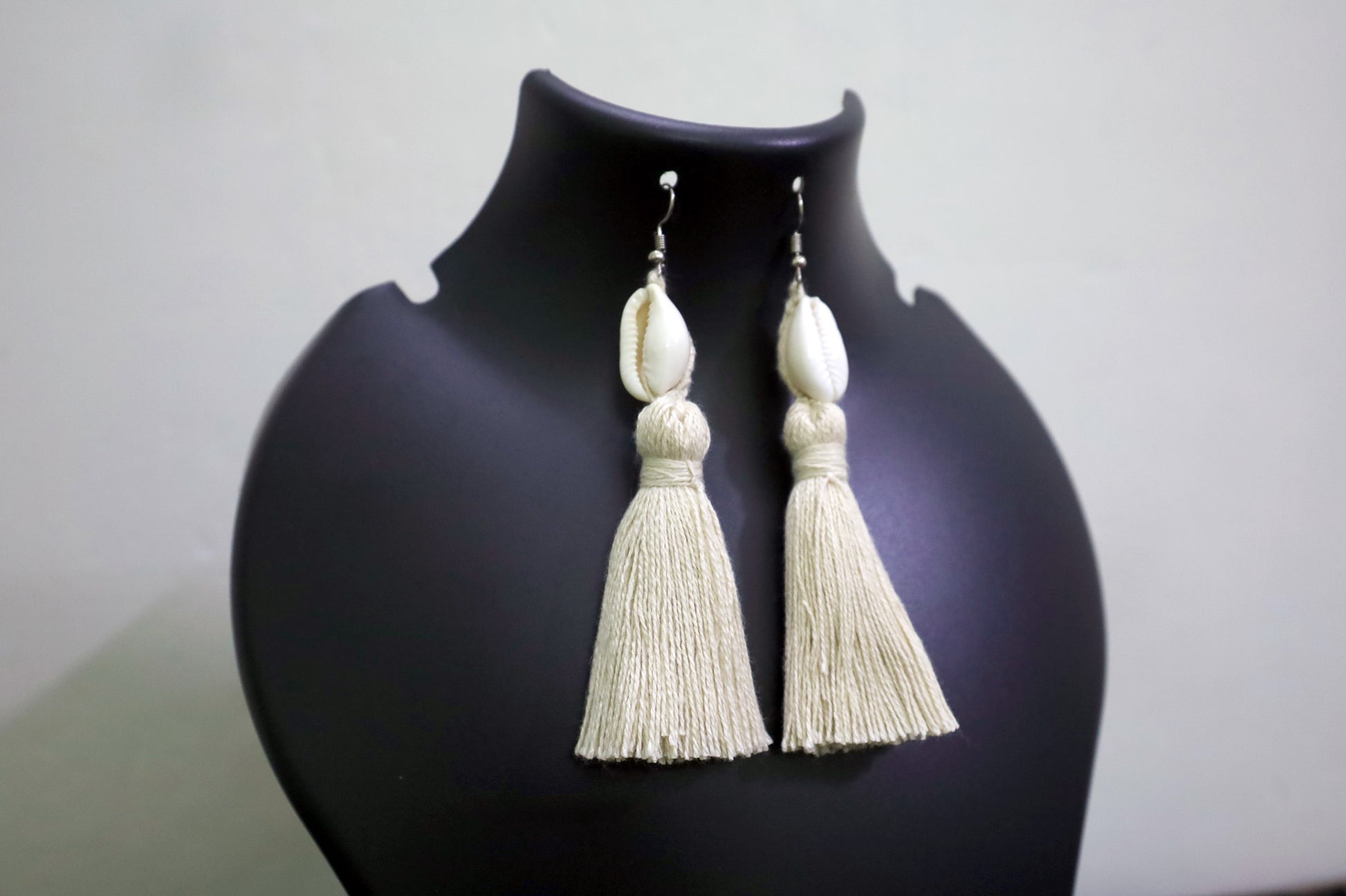 Close-up of handcrafted Cowrie Seashell & Off-white Tussle Dangle Earrings on black mannequin bust against a white background