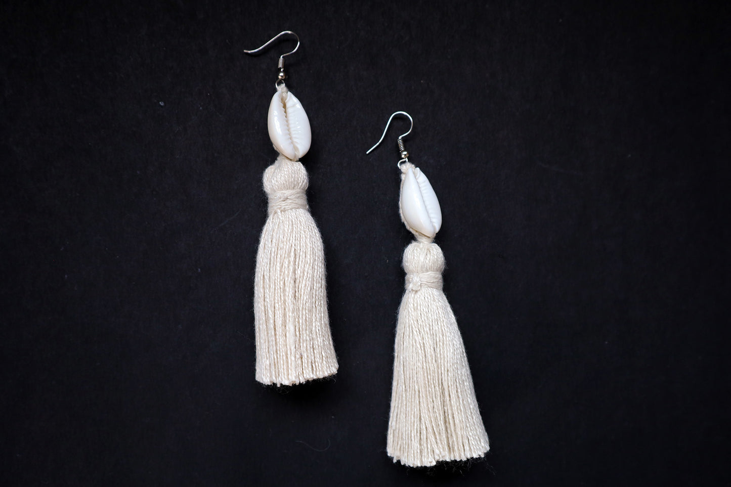 Top view of Cowrie Seashell & Off-white Tussle Dangle Earrings on black fabric background