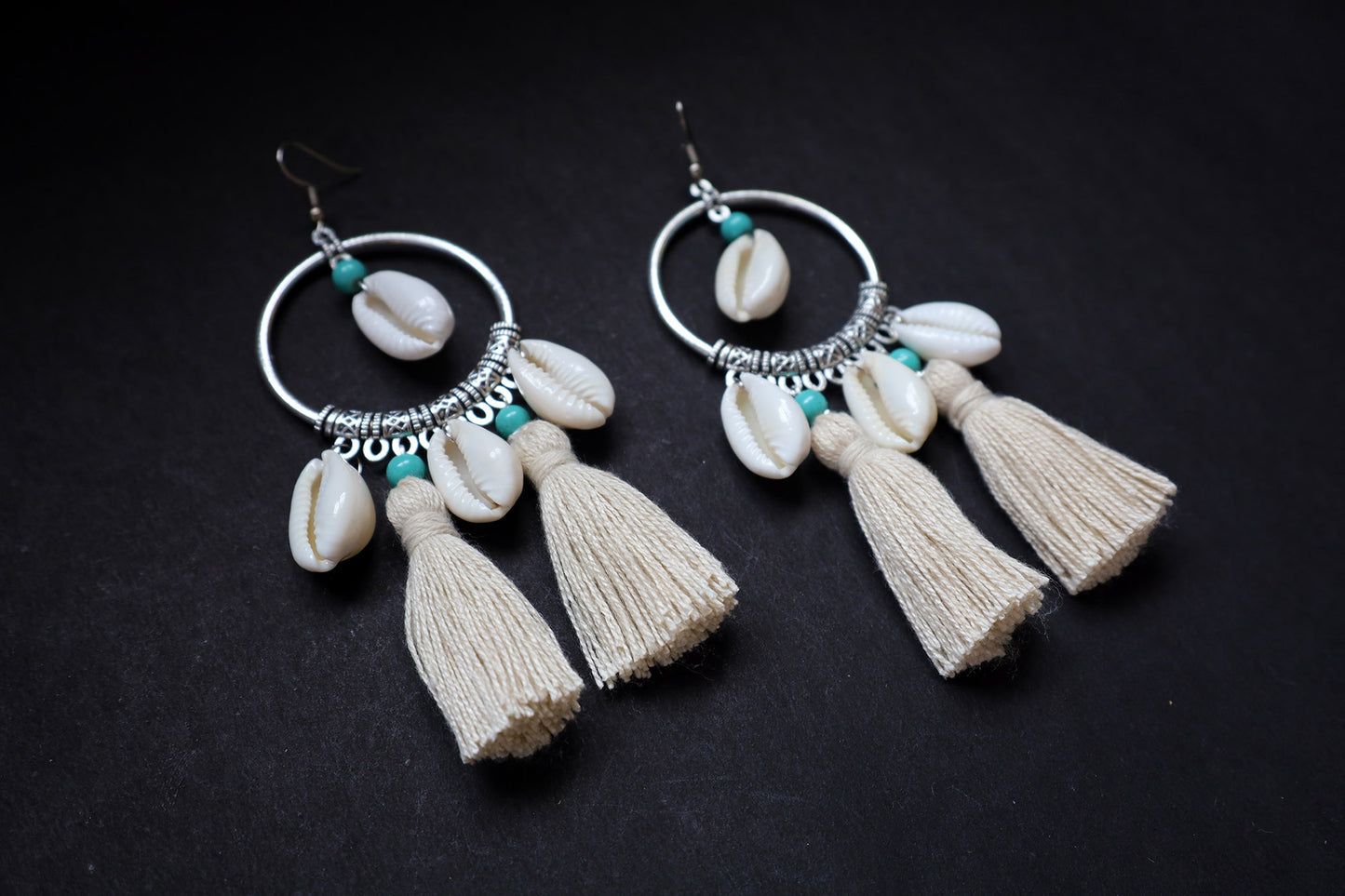 Angle view of pair of handmade cowrie seashell tussle oxidized hoop dangle earrings on fabric background