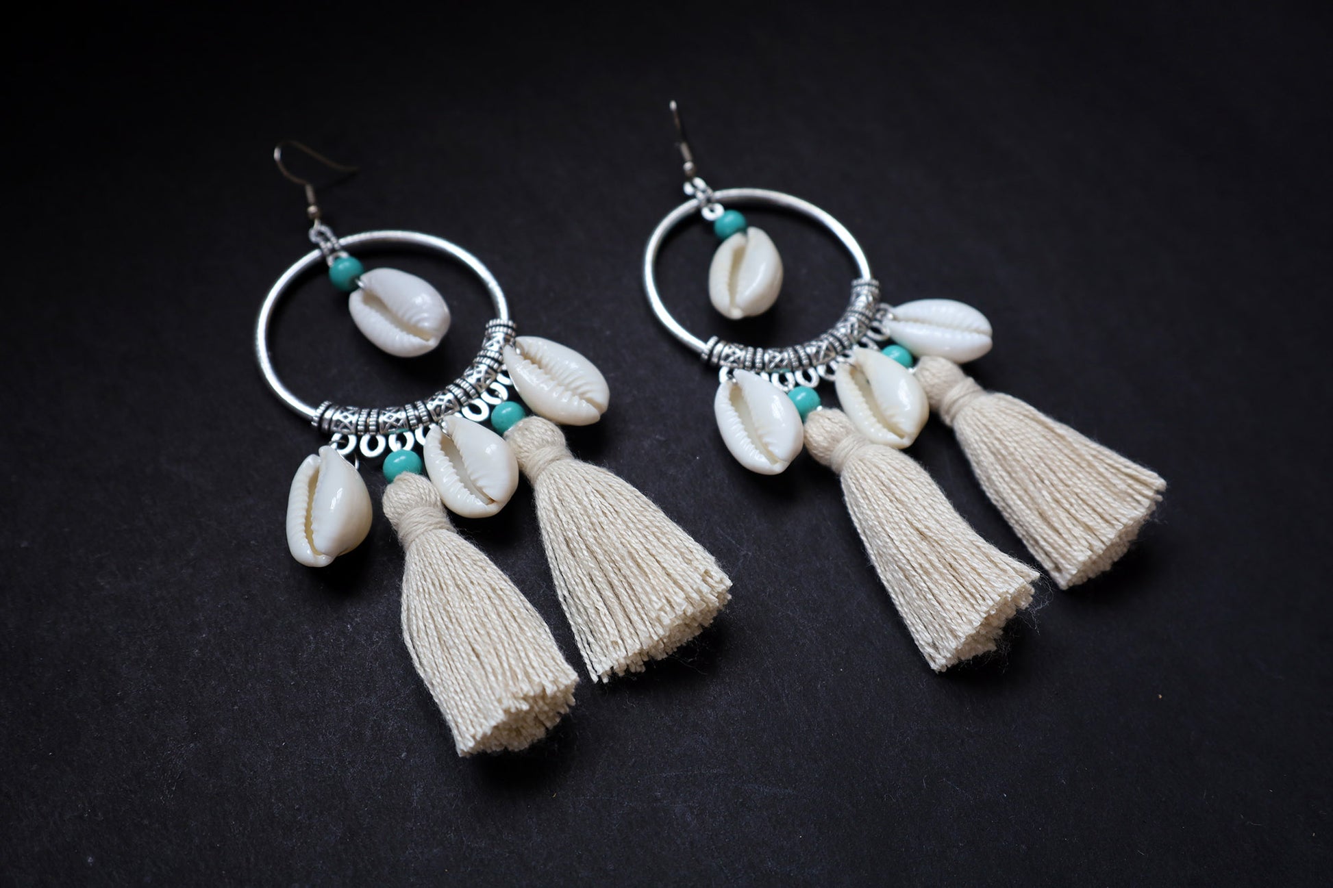Angle view of pair of handmade cowrie seashell tussle oxidized hoop dangle earrings on fabric background