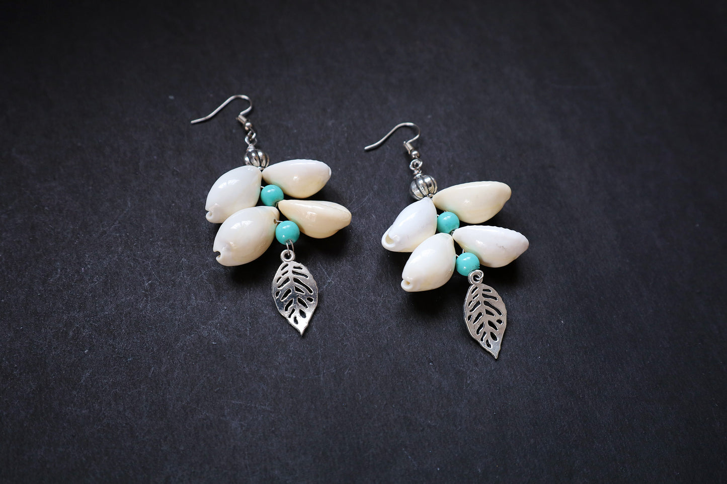 Pair of earrings with cowrie, leaf charm and bead design on a dark background