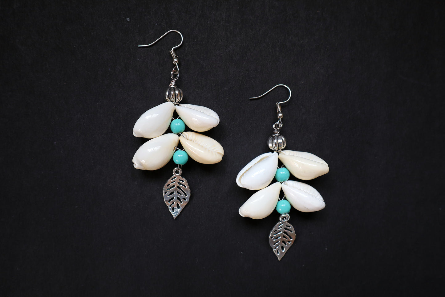 Pair of earrings with white cowrie, turquoise beads, and silver leaf charms on a black background