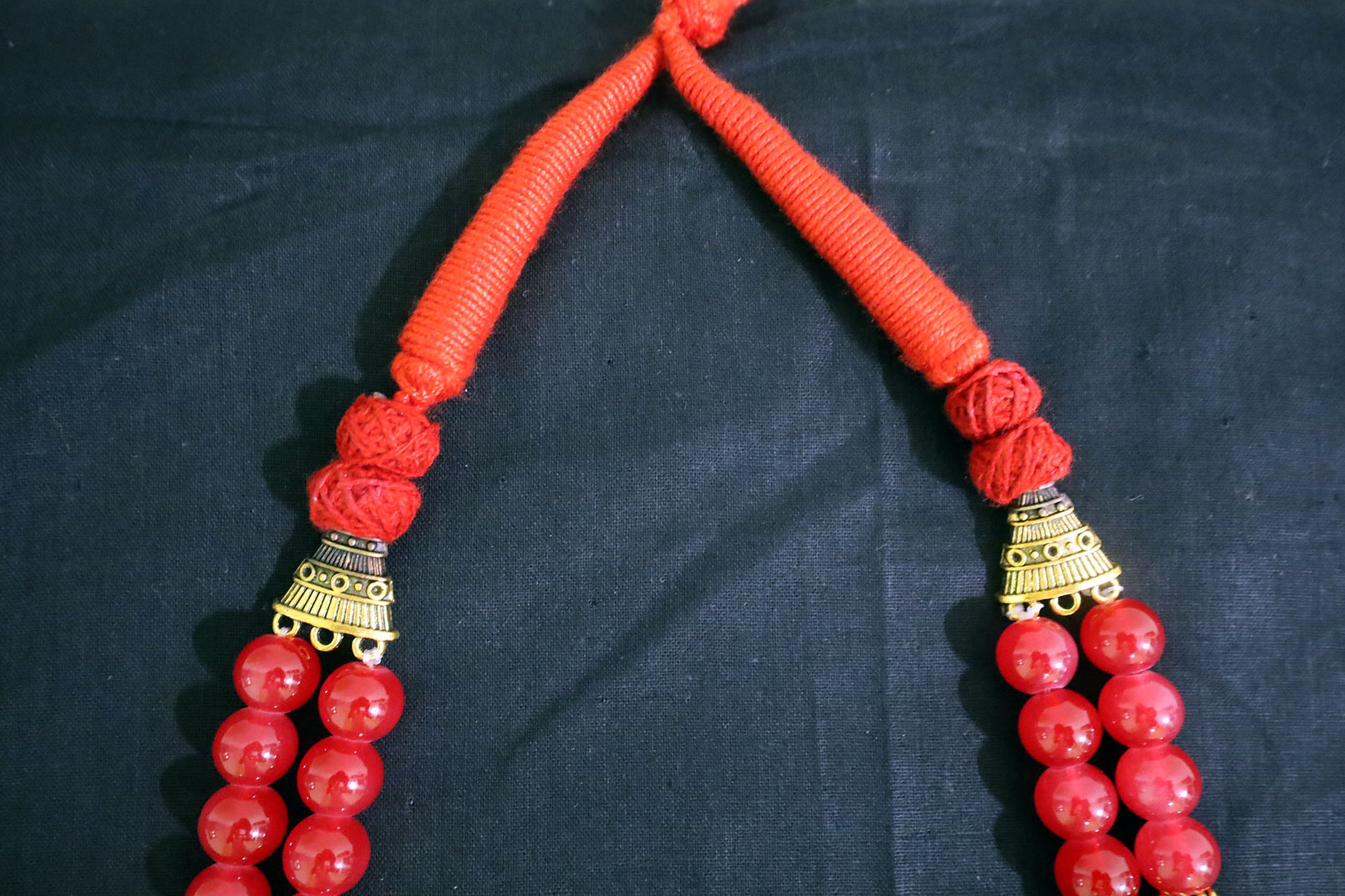 Closer top section of red necklace with glass beads and golden drop pendant on black fabric.