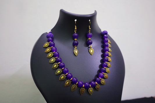 Violet glass beaded with golden leaf charm necklace and earrings set on a black mannequin bust against a white background